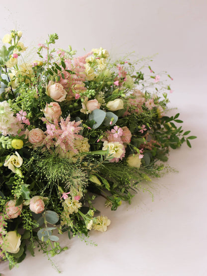 A side view of a 3ft coffin spray featuring pastel pink, yellow and peach flowers and foliage. The funeral arrangement is sitting against a neutral background and features Roses, Astlibe, Stocks and Lisianthus. 