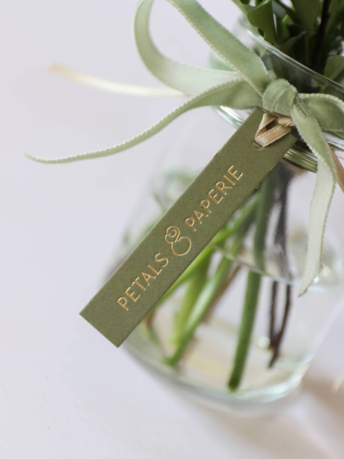 Glass vase of flowers with a green tag and gold foiled 'Petals & Paperie' logo, tied to the vase with a green velvet ribbon.
