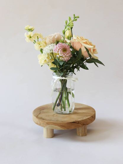 A single jam jar with pastel coloured flowers sitting on a wooden stool against a light background
