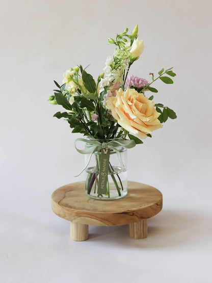 A single jam jar with pastel coloured flowers sitting on a wooden stool against a light background