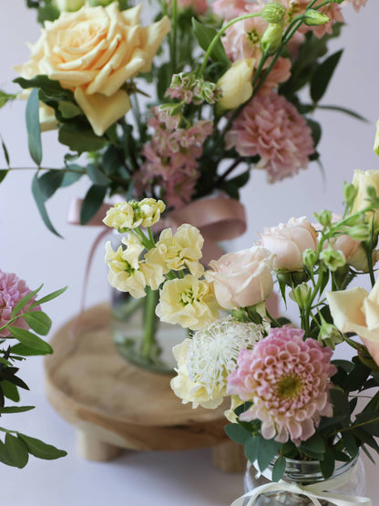 A group of pastel flowers in jam jars on a white surface with a wooden stand.
