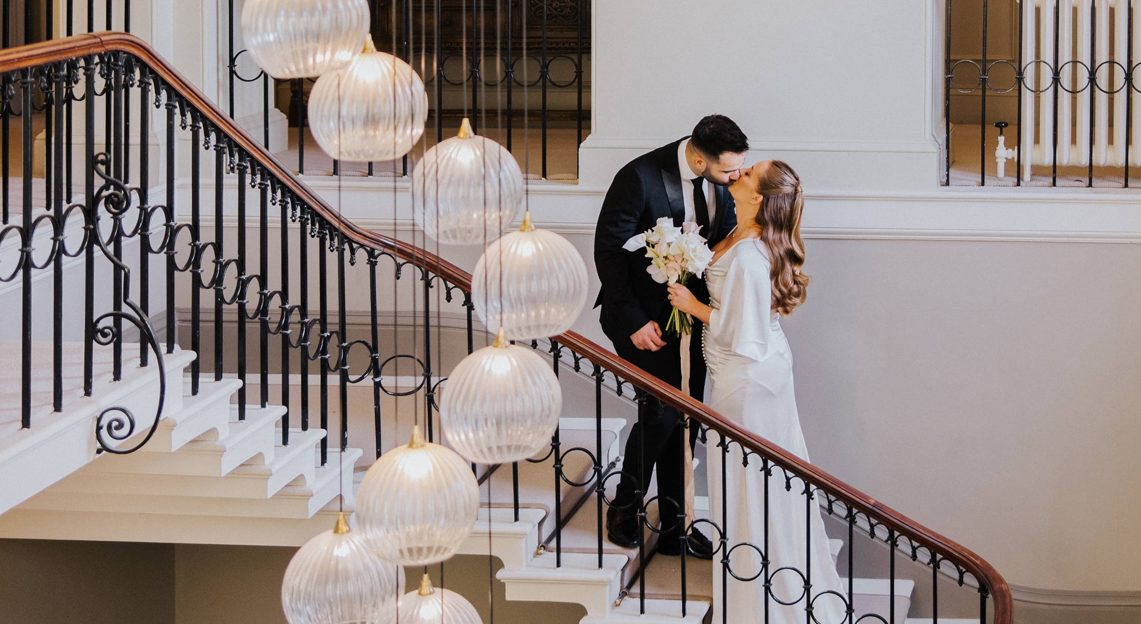 A bride wearing a silk wedding dress carrying a white and blush colour bridal bouquet and a groom in a black suit standing on the staircase at Saltmarshe Hall with a hanging chandelier. 