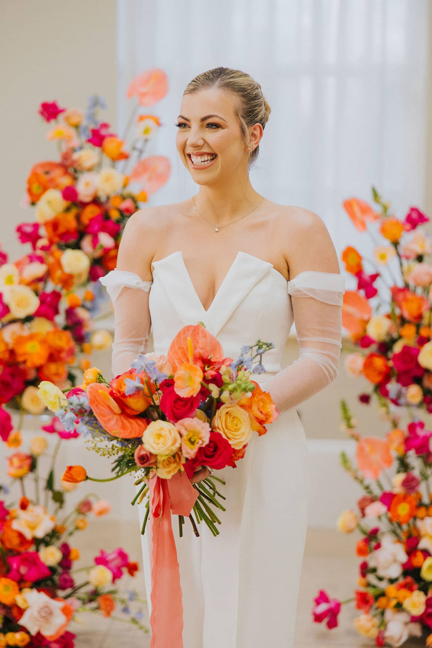 A bride wearing a white bridal jumpsuit with silk chiffon sleeves holding a colourful bouquet, stood in front of a bright and colourful pink, coral and yellow floral arch at Saltmarshe Hall.