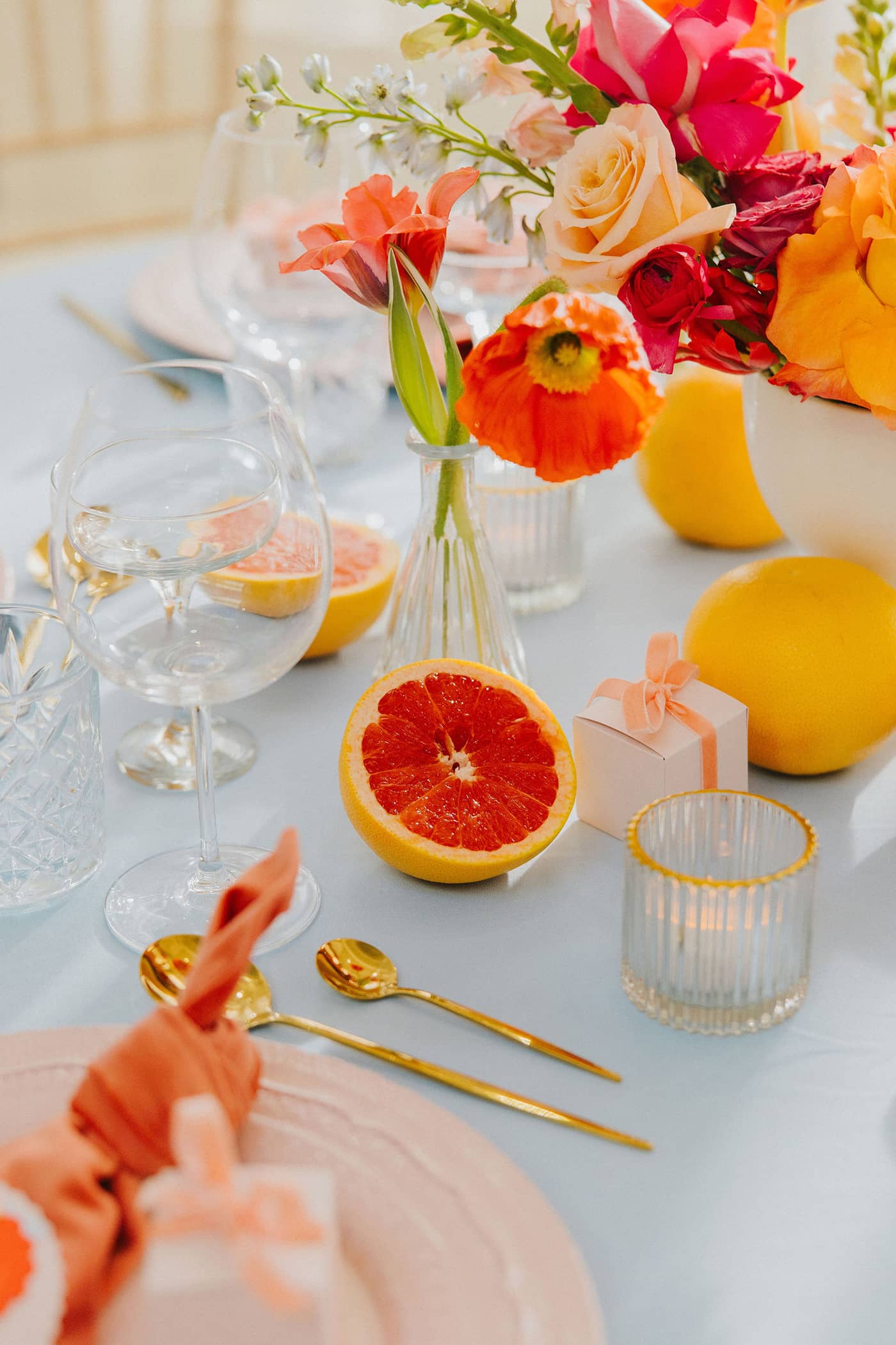 A wedding table setting with pastel pink charger plates, pastel pink favour boxes with a coral velvet ribbon and decorative pink grapefruit slices, all sitting on a pastel blue tablecloth with gold cutlery.