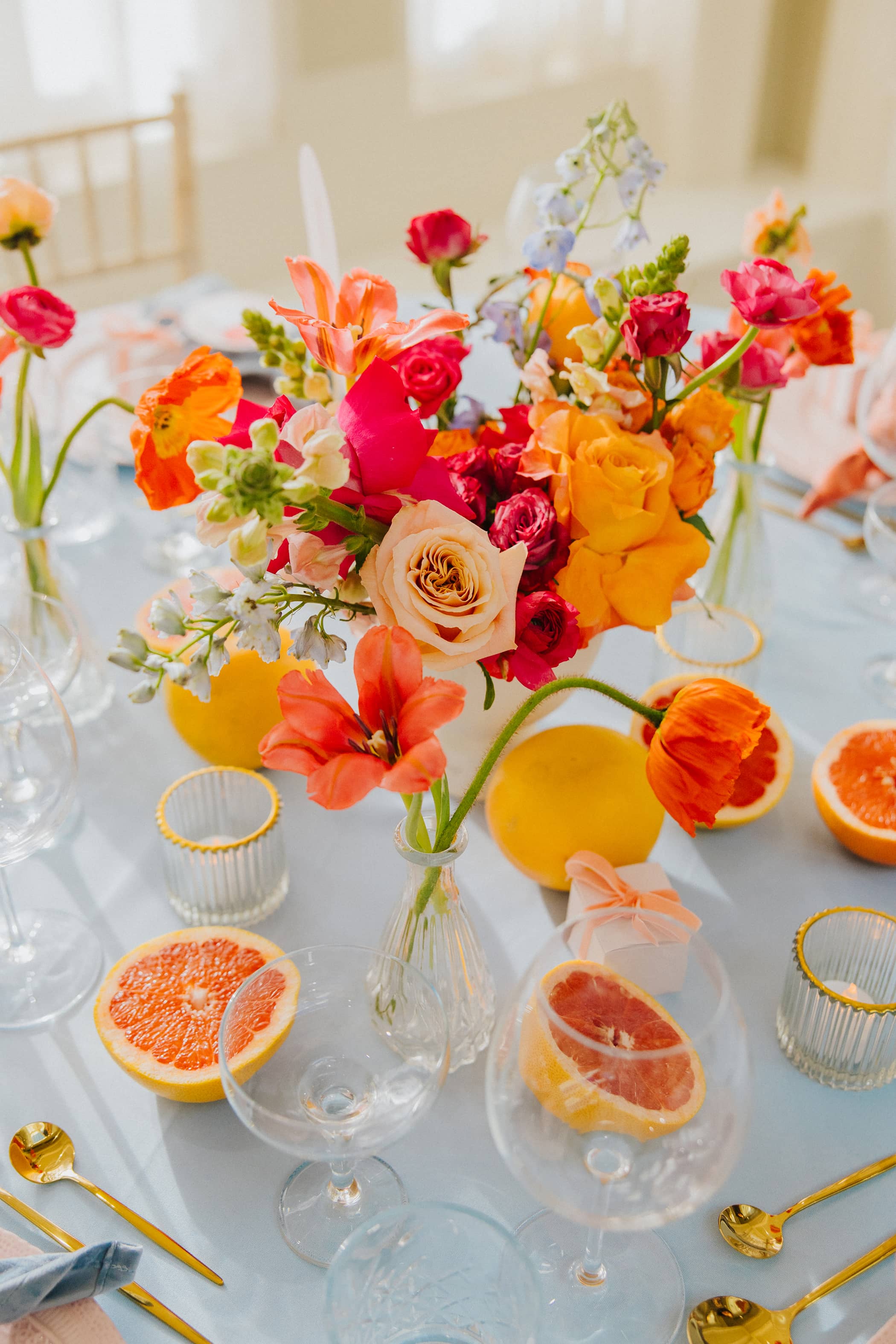 A wedding table setting with a coral, pink and blue floral arrangement and bud vases. The table also features pastel pink favour boxes with a coral velvet ribbon and decorative pink grapefruit slices, all sitting on a pastel blue tablecloth with gold cutlery.