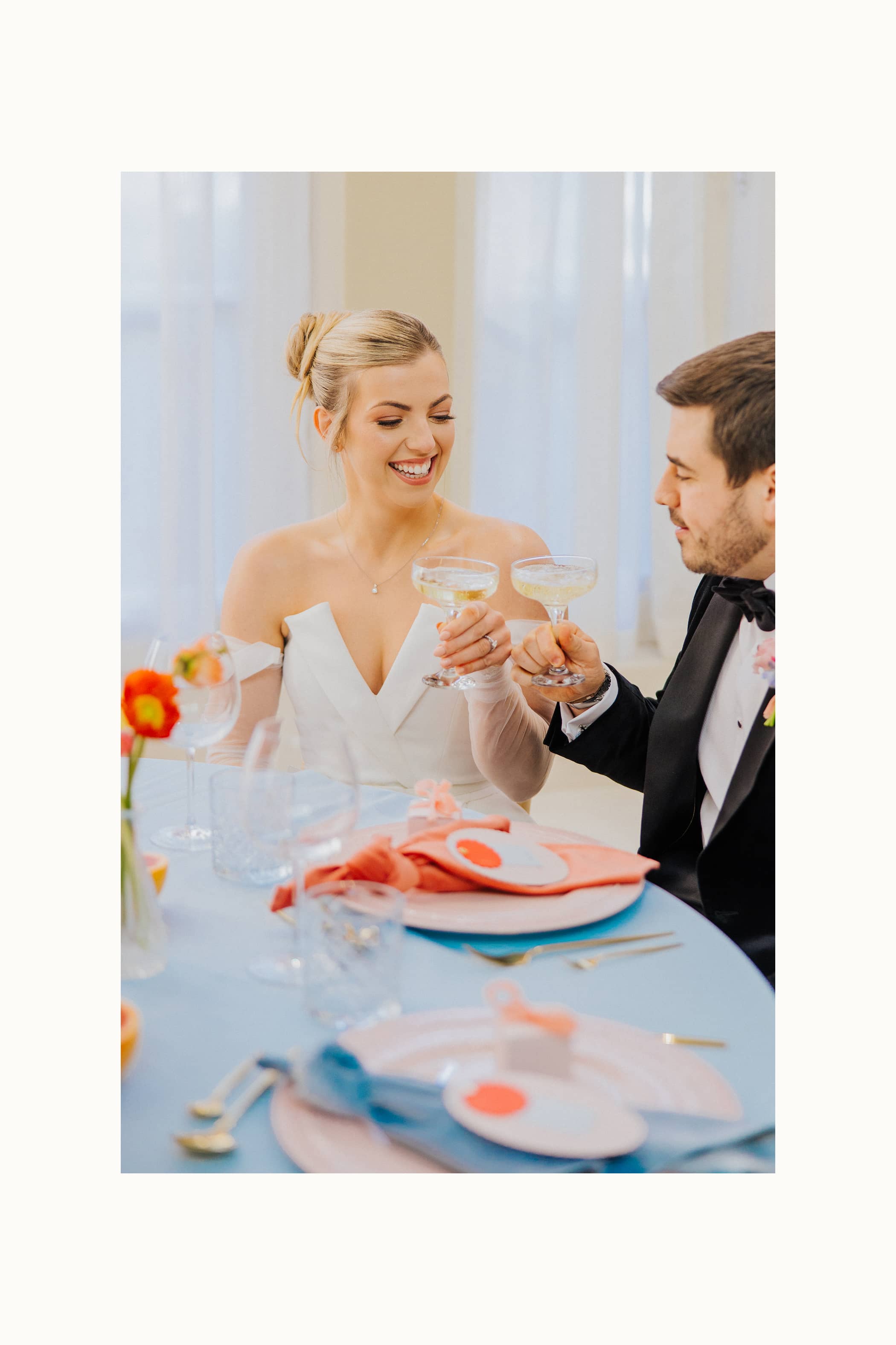 A bride dressed in a white jumpsuit and a groom in a black tuxedo sitting at a round wedding table at Saltmarshe Hall, raising two glasses of champagne in coupé glasses. The table is set with pastel pink charger plates on a pastel blue table cloth.