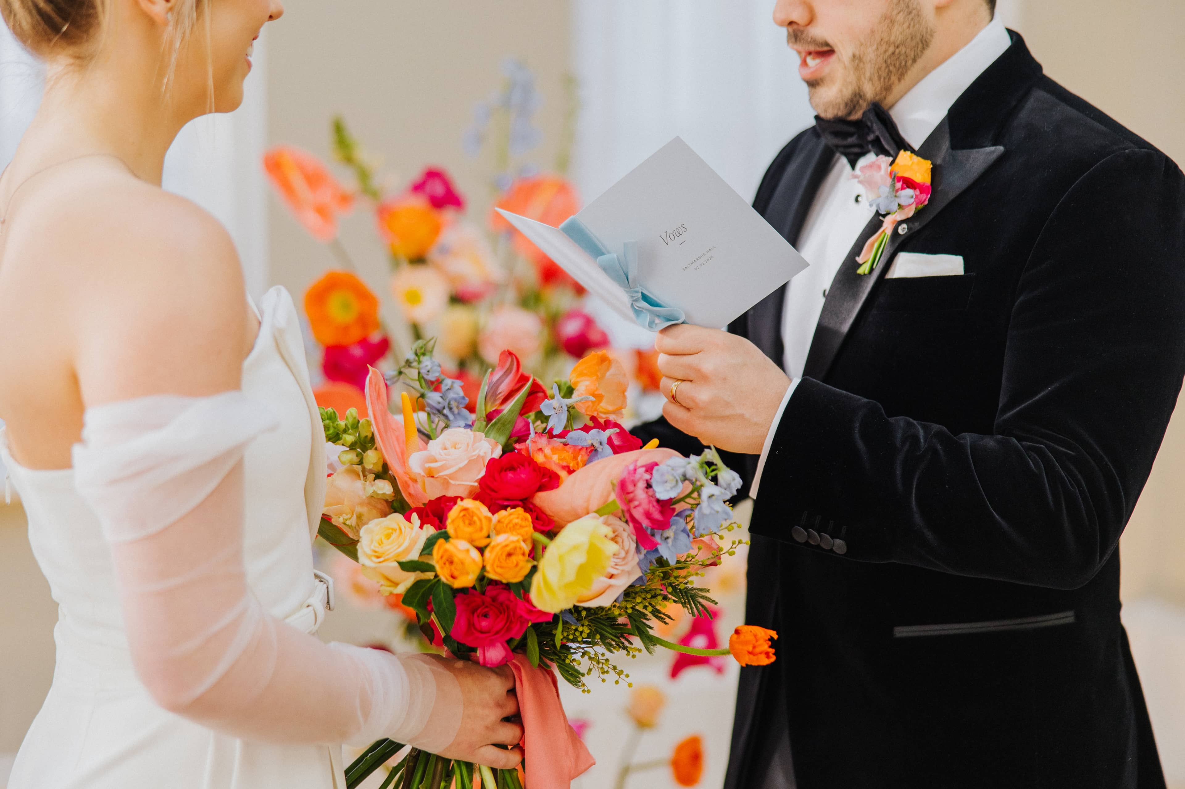 A close up of a bride dressed in a white jumpsuit and a groom in a black tuxedo exchanging vows in front of a bright and vibrant floral arch at Saltmarshe Hall. The bride is carrying a bright and colourful wedding bouquet and the groom is holding a pastel blue vow book.