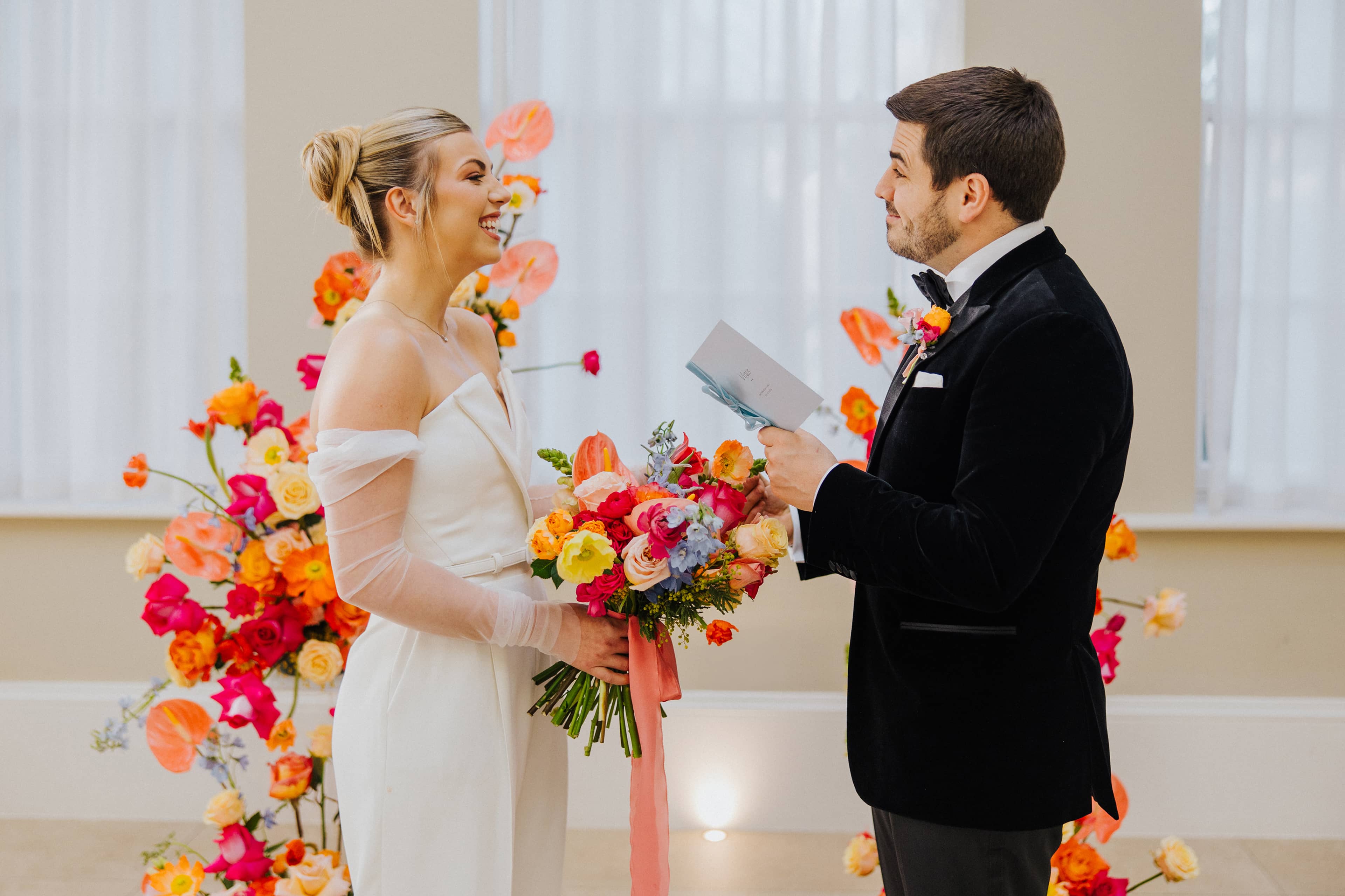 A bride dressed in a white jumpsuit and a groom in a black tuxedo exchanging vows in front of a bright and vibrant floral arch at Saltmarshe Hall. The bride is carrying a bright and colourful wedding bouquet and the groom is holding a pastel blue vow book.