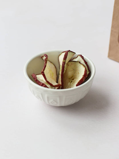 5 dried red apple slices for Christmas wreath making, positioned in a cream decorative bowl, on a light background. There is a bag of dried apple slices in a brown kraft bag in the background.