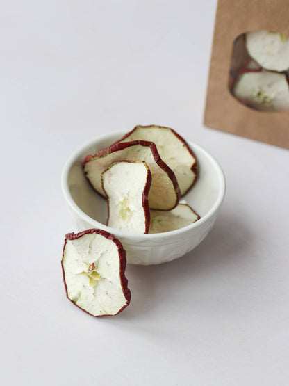 5 dried red apple slices for Christmas wreath making, positioned in a cream decorative bowl, on a light background. There is a bag of dried apple slices in a brown kraft bag in the background.