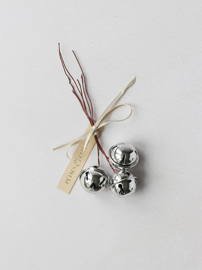 3 wired silver jingle bells, sitting on a light grey background. The bells are tied together with a Petals & Paperie cream gift tag and cream satin ribbon.