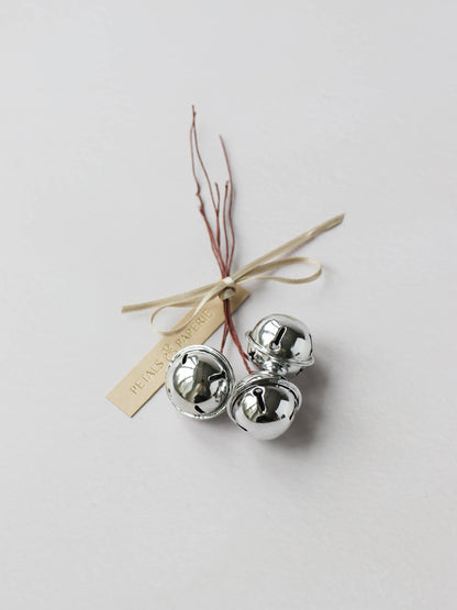 3 wired silver jingle bells, sitting on a light grey background. The bells are tied together with a Petals & Paperie cream gift tag and cream satin ribbon.