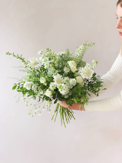 A woman holding a white and green bouquet of flowers out to the side against a neutral background. The bouquet includes Delphiniums, Stocks, Roses and Ammi.