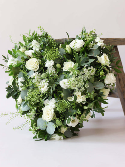 Heart shaped funeral posy with white and green flowers on a white background leaning against a wooden stool