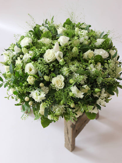 Round funeral posy with white and green flowers on a white background sitting on a wooden stool