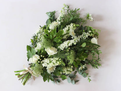 A top down view of a white and green tied funeral sheaf containing white flowers and green foliages, tied with a champagne colour satin ribbon. The sheaf contains Delphiniums, Snapdragons, Ammi and Roses.