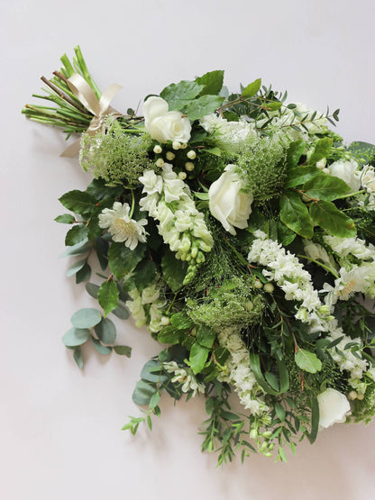 A top down view of a white and green tied funeral sheaf containing white flowers and green foliages, tied with a champagne colour satin ribbon. The sheaf contains Delphiniums, Snapdragons, Ammi and Roses.