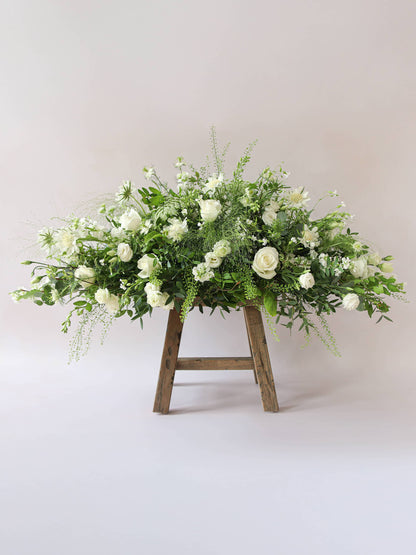 A side view of a 3ft coffin spray featuring white and green flowers and foliage. The funeral arrangement is sitting on a rustic wooden stool against a neutral background and features white Roses, Ammi, Stocks and Lisianthus. 