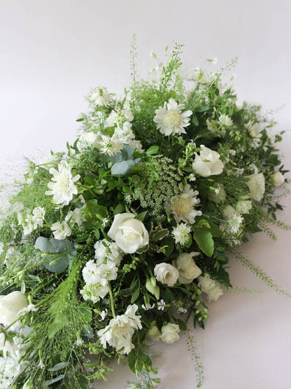 A diagonal top view of a 3ft coffin spray featuring white and green flowers and foliage. The funeral arrangement is sitting against a neutral background and features white Roses, Ammi, Stocks and Lisianthus. 