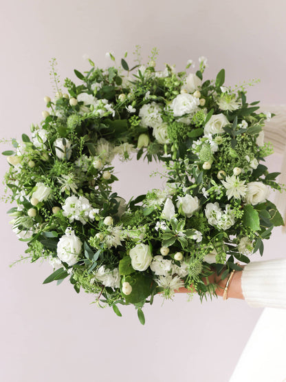 Funeral wreath with white and green flowers being held by a women in neutral clothing against a white background