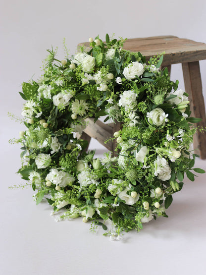 Funeral wreath with white and green flowers leaning against a wooden stool against a white background
