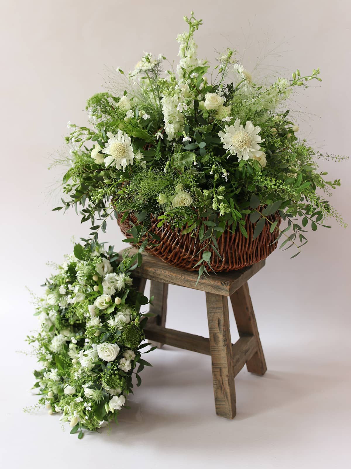 Wicker basket with a floral arrangement inside on a wooden stool against a white background, with a white and green funeral posy below. 