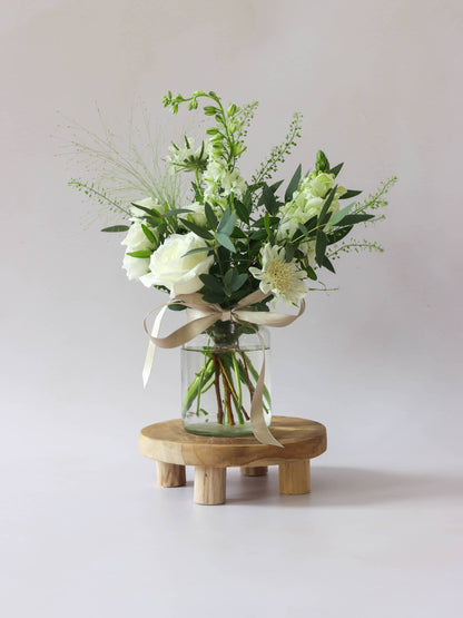 A single jam jar with white and green flowers sitting on a wooden stool against a light background