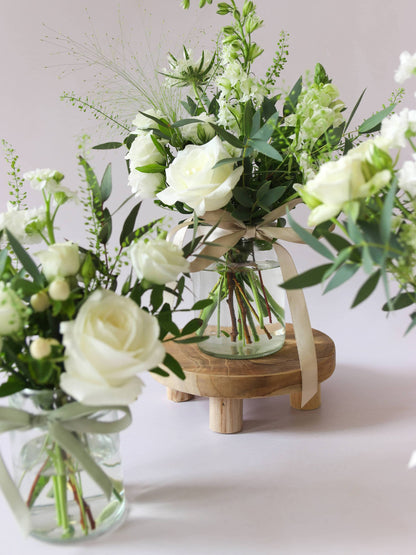 Two jam jars with white and green flowers sitting on a wooden stool against a light background