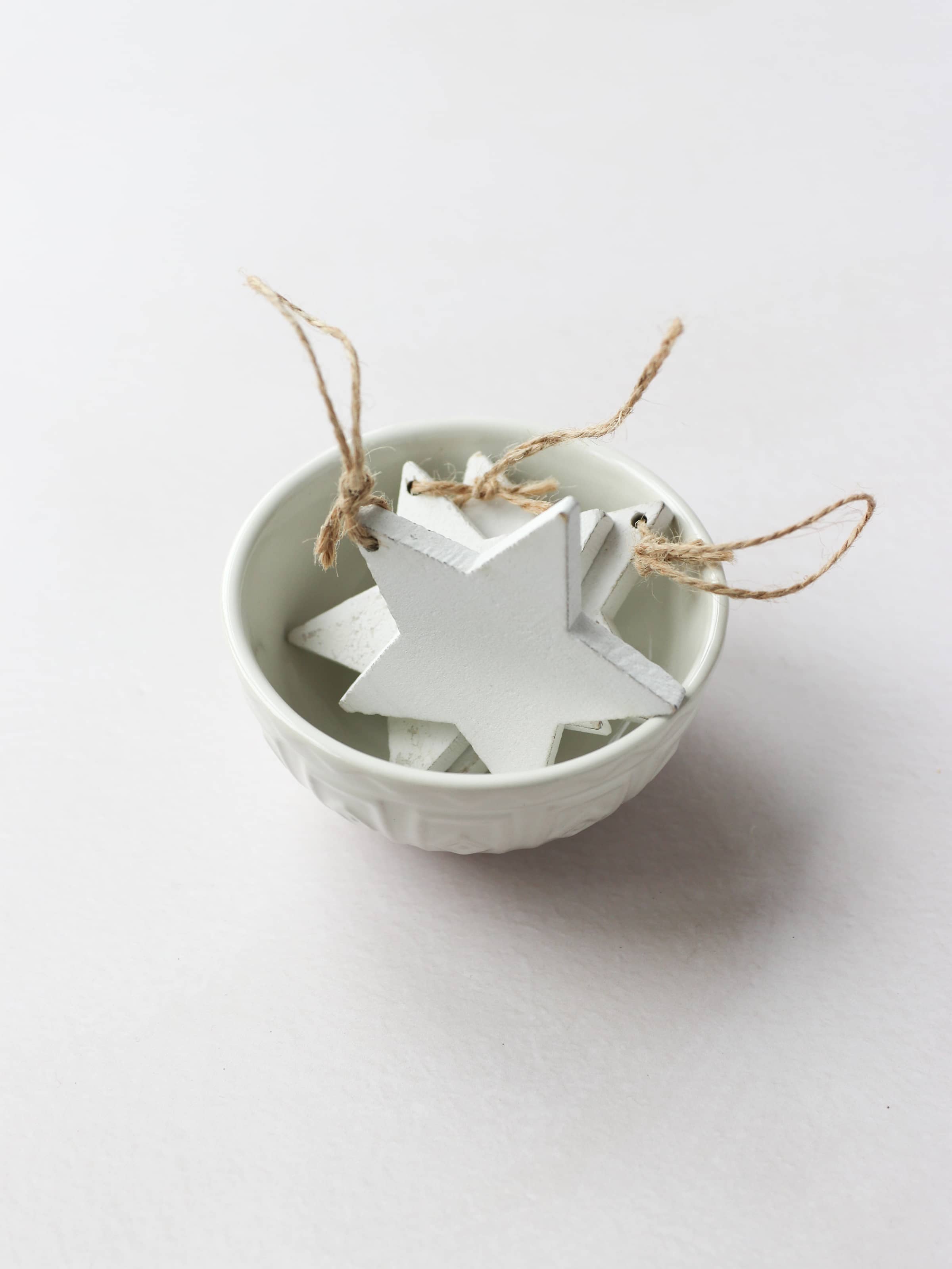 3 white star-shaped wooden ornaments for Christmas wreath making with brown twine tie loops, sitting in a cream decorative bowl on a light grey background.