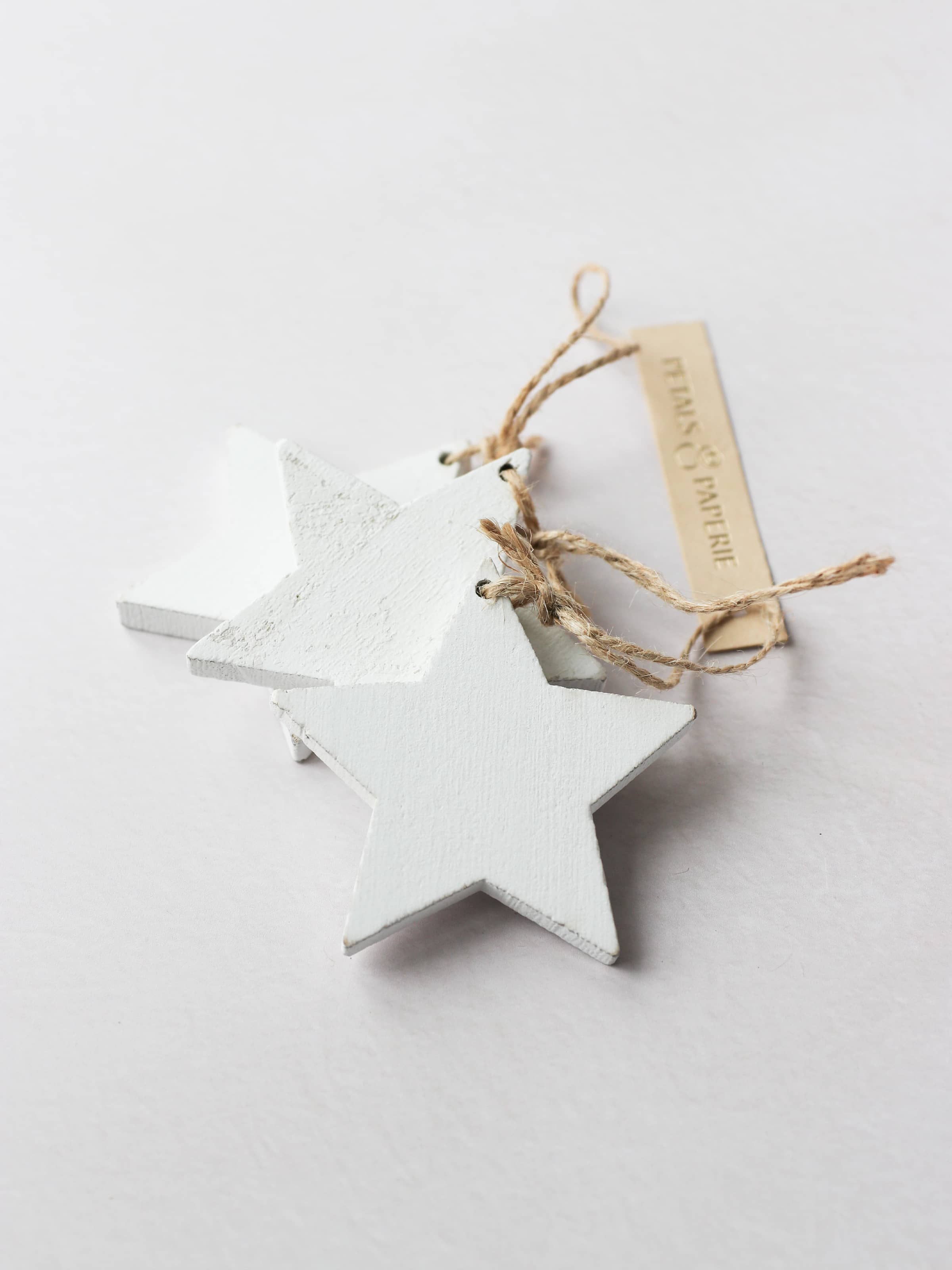 3 white star-shaped wooden ornaments for Christmas wreath making with brown twine tie loops, on a light grey background. The stars are tied together with a Petals & Paperie gift tag.