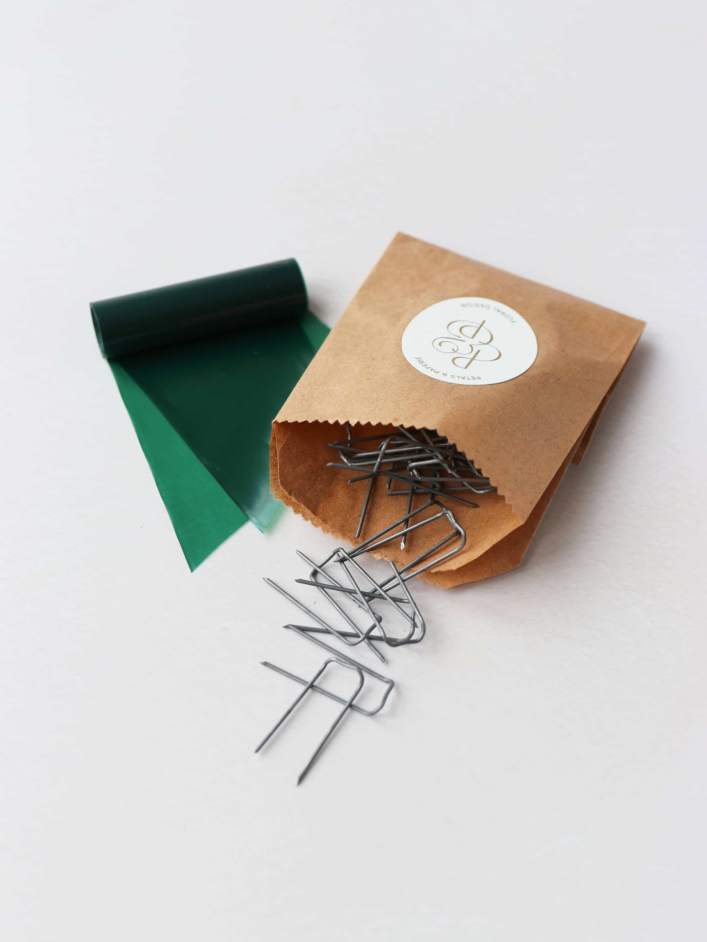 A kraft paper bag containing metal mossing pins and roll of green wreath wrap, sitting on a neutral background.
