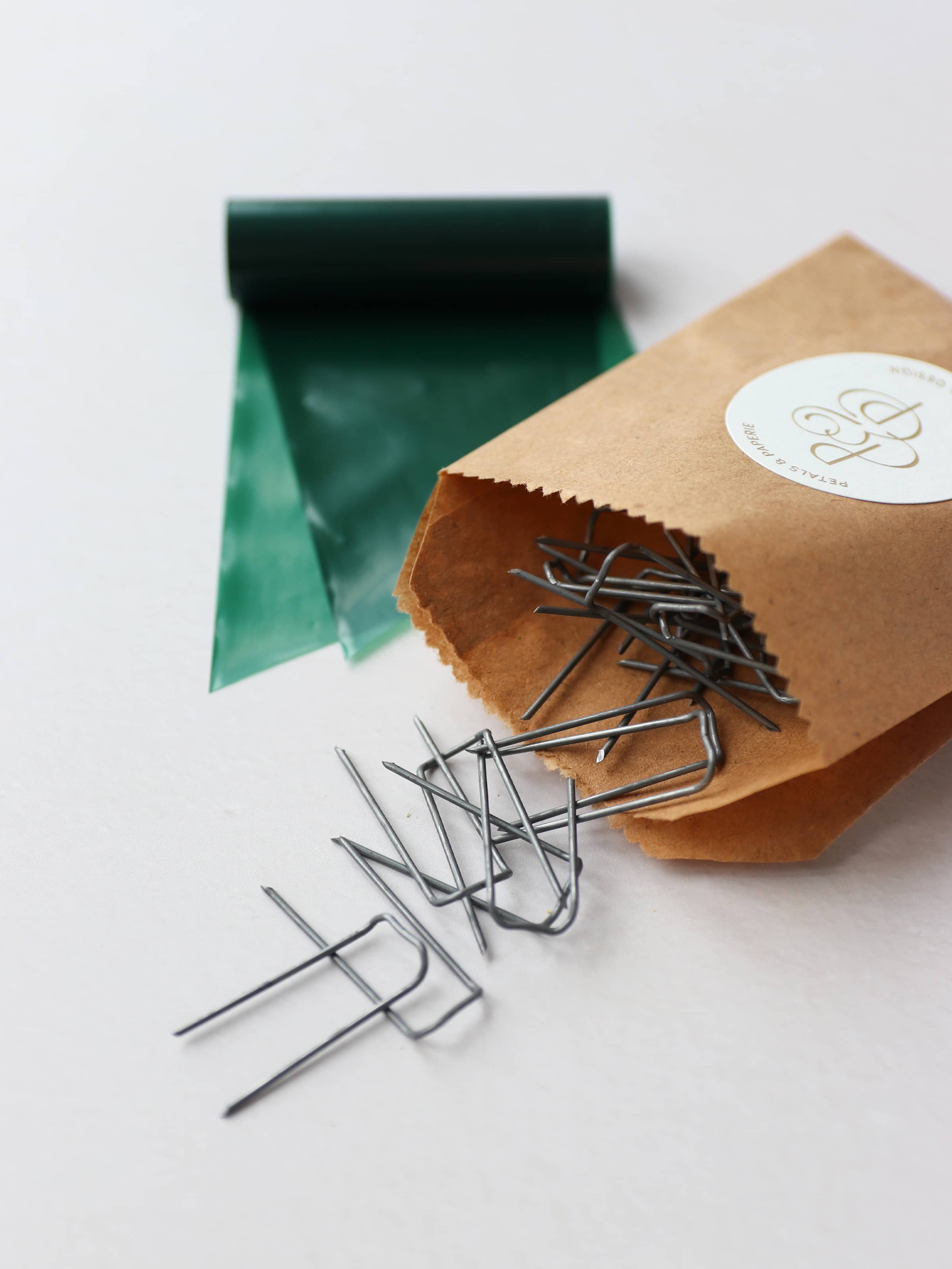 A close up of a kraft paper bag containing metal mossing pins and roll of green wreath wrap, sitting on a neutral background.